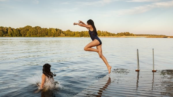 Two girls jumping into the water from a bathing jetty by a lake on a summer evening