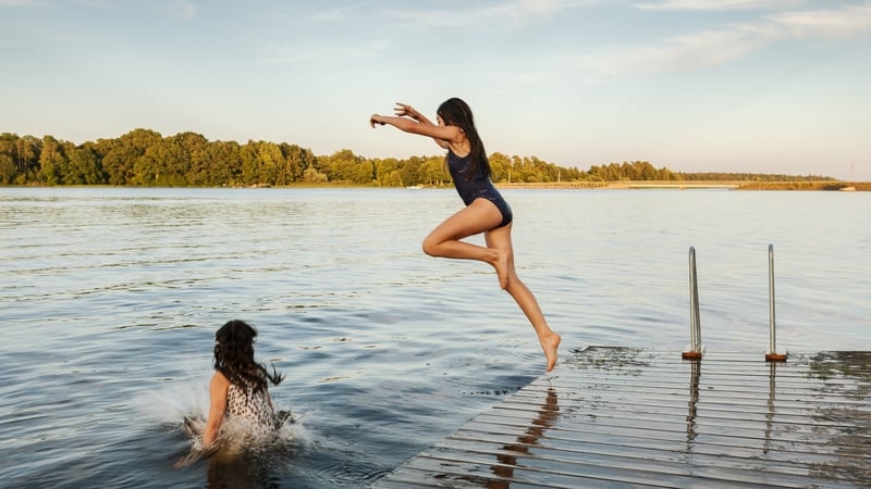 Two girls jumping into the water from a bathing jetty by a lake on a summer evening