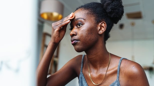 Young multiracial woman applying foundation on face with sponge while standing in front of mirror trying to hide skin problems