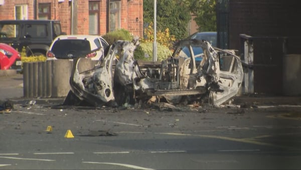 Image shows the burnt out remains of a vehicle on a road in daylight.