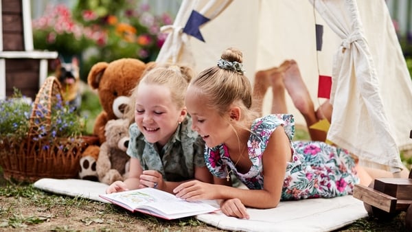 Two girls reading a book in their garden inside a wigwam with a teddy bear.