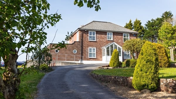 Image shows a large redbrick building with white window frames at the top of a driveway. The sky is blue and there are lots of trees in the image.