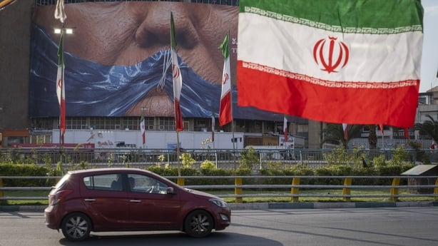 A vehicle drives through a square beneath a billboard depicting an AI-generated image of the Strait of Hormuz