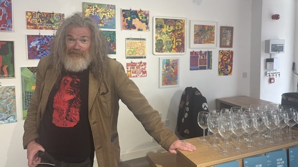 Image shows an older man with shoulder length hair and a beard standing in front of a white wall with several colourful paintings.