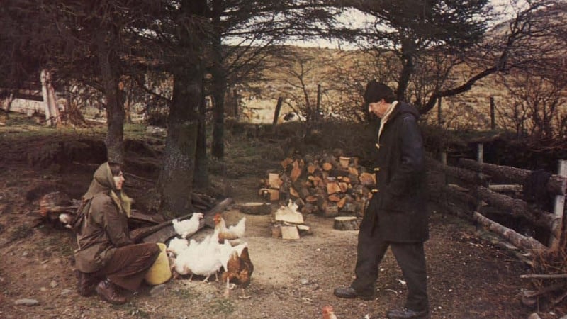 A woman in a brown coat sitting next to some chickens, observed by a man in a dark coat and hat