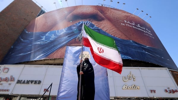A woman holds the Iranian flag as she stands in front of an anti-US billboard referring to President Donald Trump and the Strait of Hormuz