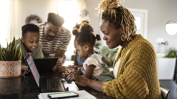 Mother working from home while holding toddler, family in background