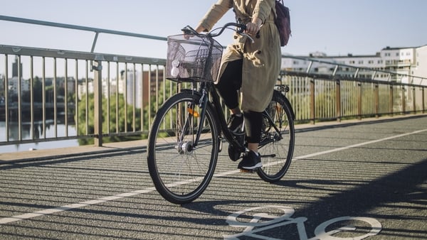 Image shows a woman with hair tied back and wearing a long coat cycling through a city