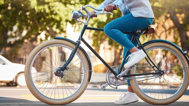 Image shows a woman wearing blue jeans cycling in a sunlight area.