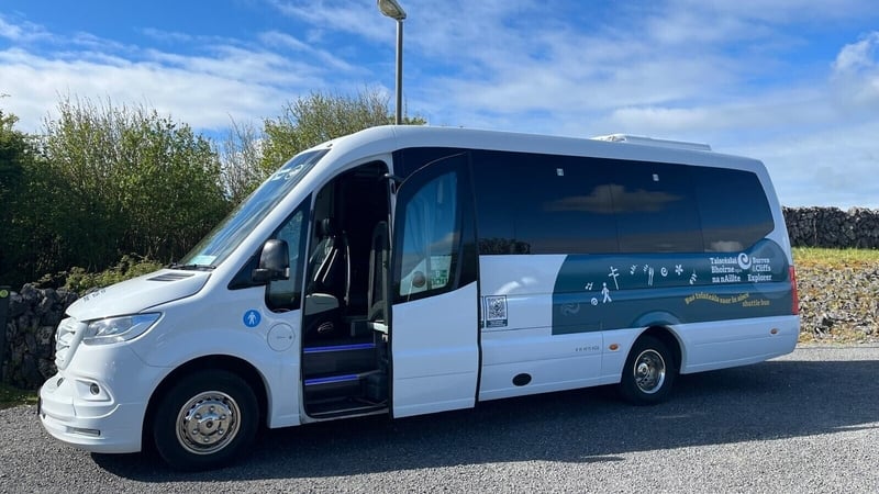 Image shows a medium sized white bus parked at the side of a road. There are trees and a blue sky in the background.