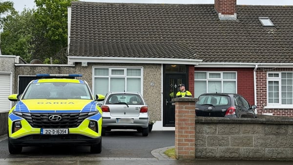 a garda car is seen in front of a bungalow with a garda in the doorway of the house and two cars parked in the driveway