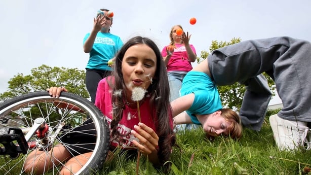 A young girl blows a dandelion in the foreground as youngster juggle and perform a back flip in the backlground at the Cruinniú na nÓg 2026 launch at TUD Grangegorman