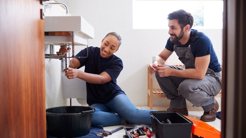 Male Plumber Teaching Female Apprentice To Fix Leaking Sink In Home Bathroom