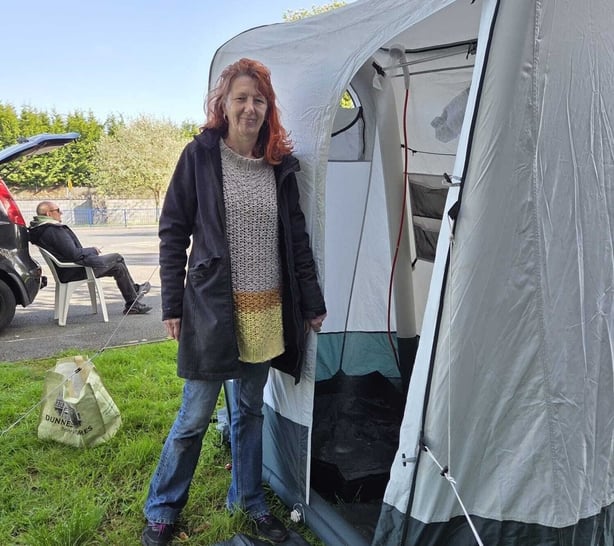 Image shows a smiling red haired woman wearing blue jeans, a beige jumper and a dark jacket. She is standing beside the entrance to a tent that contains a mobile shower unit.