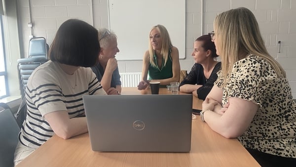 A number of people are pictured at a desk, talking