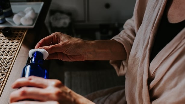Close-up image of a woman holding a bottle containing liquid, and dampening a cotton ball in preparation to cleanse, tone and moisturise her skin.