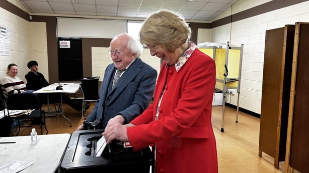 michael d higgins and sabina higgins put a ballot paper into a ballot box as two people watch on