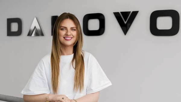 Katelyn Miller wears a white t shirt as she poses in front of a white wall and black Damovo sign
