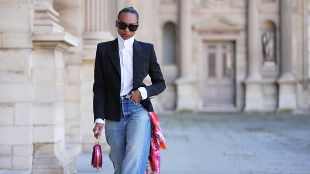 PARIS, FRANCE - APRIL 24: Emilie Joseph wears black hair in a bun, black sunglasses from Gentle Monster, a multicolor silk scarf by Kenzo, a black structured blazer jacket by Balmain, a white mandarin-collar shirt by Figaret Paris, blue wide-leg jeans by Saint Laurent with hems, a pink top-handle ba