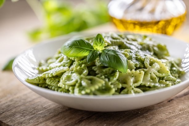 A plate of farfalle pasta, richly coated in fresh basil pesto and garnished with a basil leaf, complemented by the basic ingredients of the sauce, such as aromatic garlic, parmesan, quality olive oil and fresh herbs in the background.